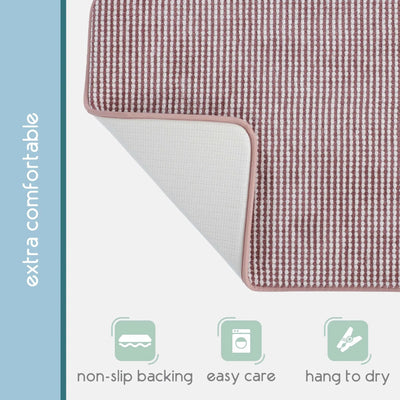 A close-up of a rectangular berry color bath rug. A vertical blue-gray sidebar reads 'extra comfortable,' and three pale green icons at the bottom indicate non-slip backing, easy care, and hang-to-dry capability.