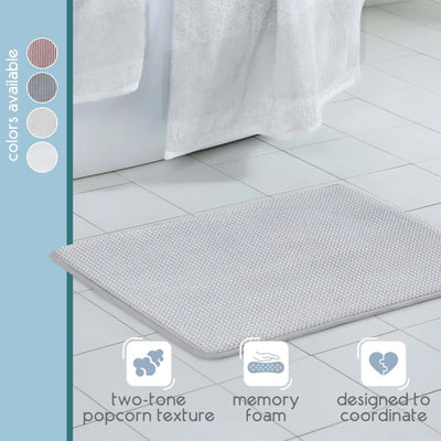 A light gray bath rug with a textured popcorn surface, shown on a white tiled bathroom floor. A vertical sidebar on the left lists available colors, and three feature icons at the bottom mention two-tone popcorn texture, memory foam, and designed to coordinate.
