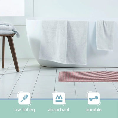 A white freestanding bathtub in a bright bathroom setting with white towels draped over the tub, a berry color bath rug on the floor, and three feature icons at the bottom reading low-linting, absorbent, and durable.