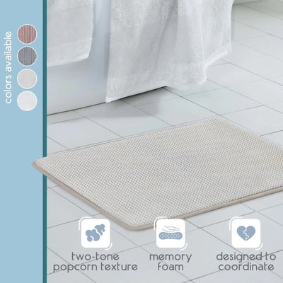 A sand color textured bath rug shown on a white tiled bathroom floor near a bathtub with a white towel. A vertical sidebar on the left reads 'colors available' with circular swatches, and three feature icons at the bottom mention 'two-tone popcorn texture,' 'memory foam,' and 'designed to coordinate.
