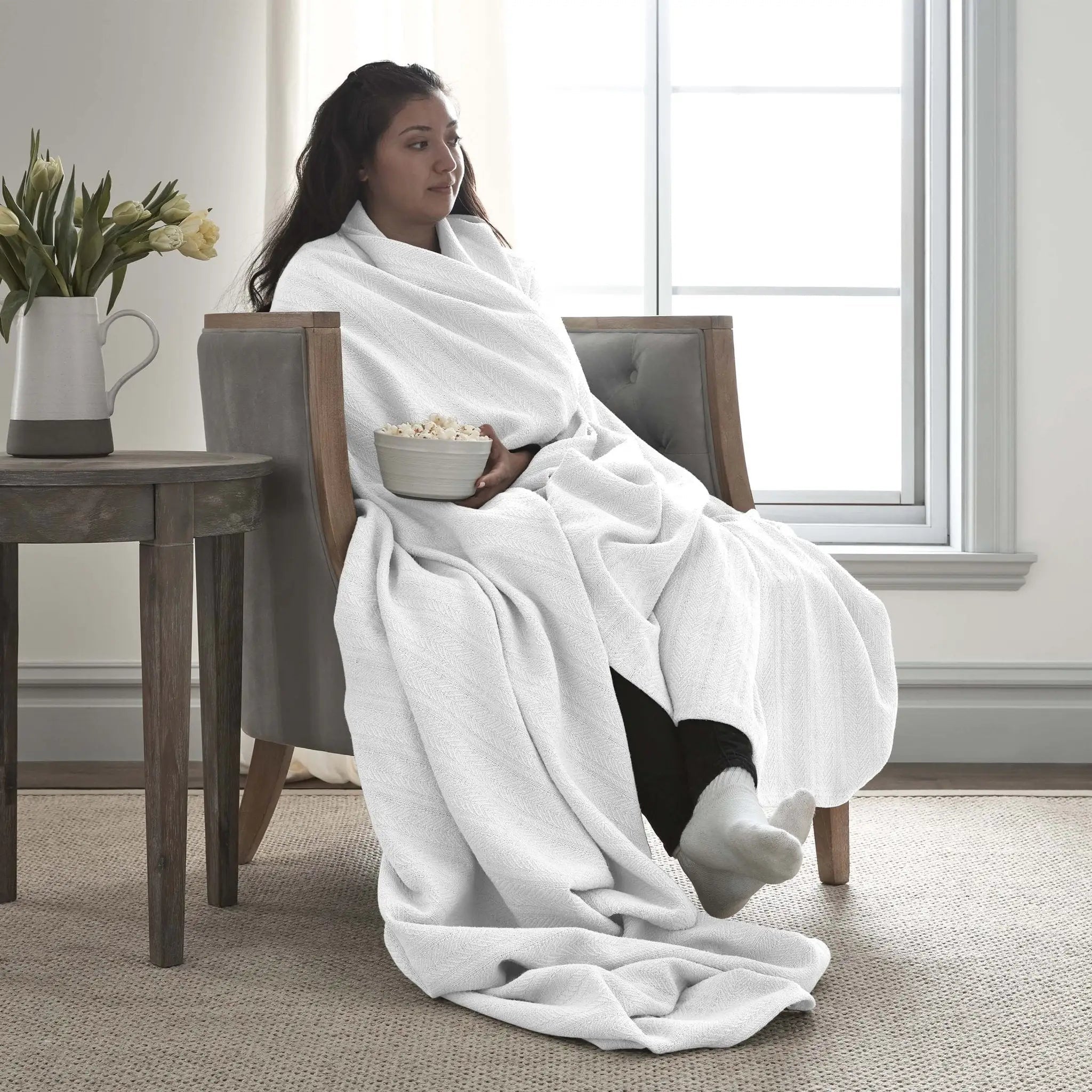 A woman sits in a gray armchair wrapped in a white textured blanket or throw, holding a bowl of popcorn in a bright living room near a window.