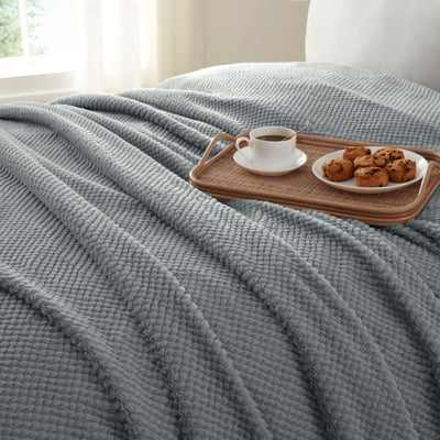 Gray textured blanket draped over a bed with a tray of coffee and pastries.
