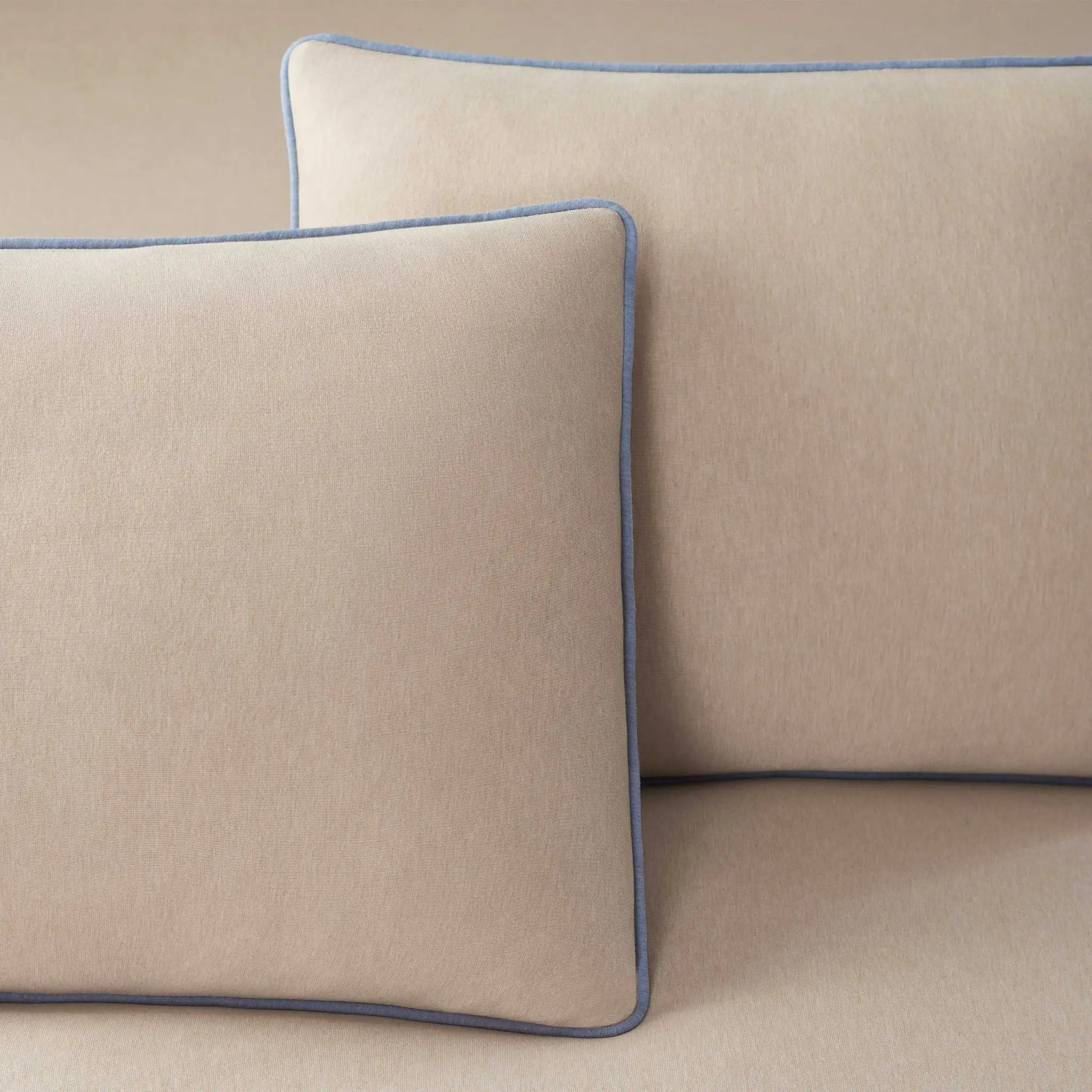 Two square beige throw pillows with blue piping trim, arranged on a matching beige fabric surface.