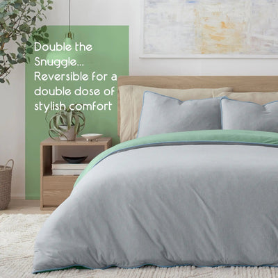 A double bed dressed in a light gray reversible comforter set and matching pillow shams with a mint green edge trim, styled in a bedroom with a wooden headboard, nightstand, wall art, and a green accent wall panel with product text.
