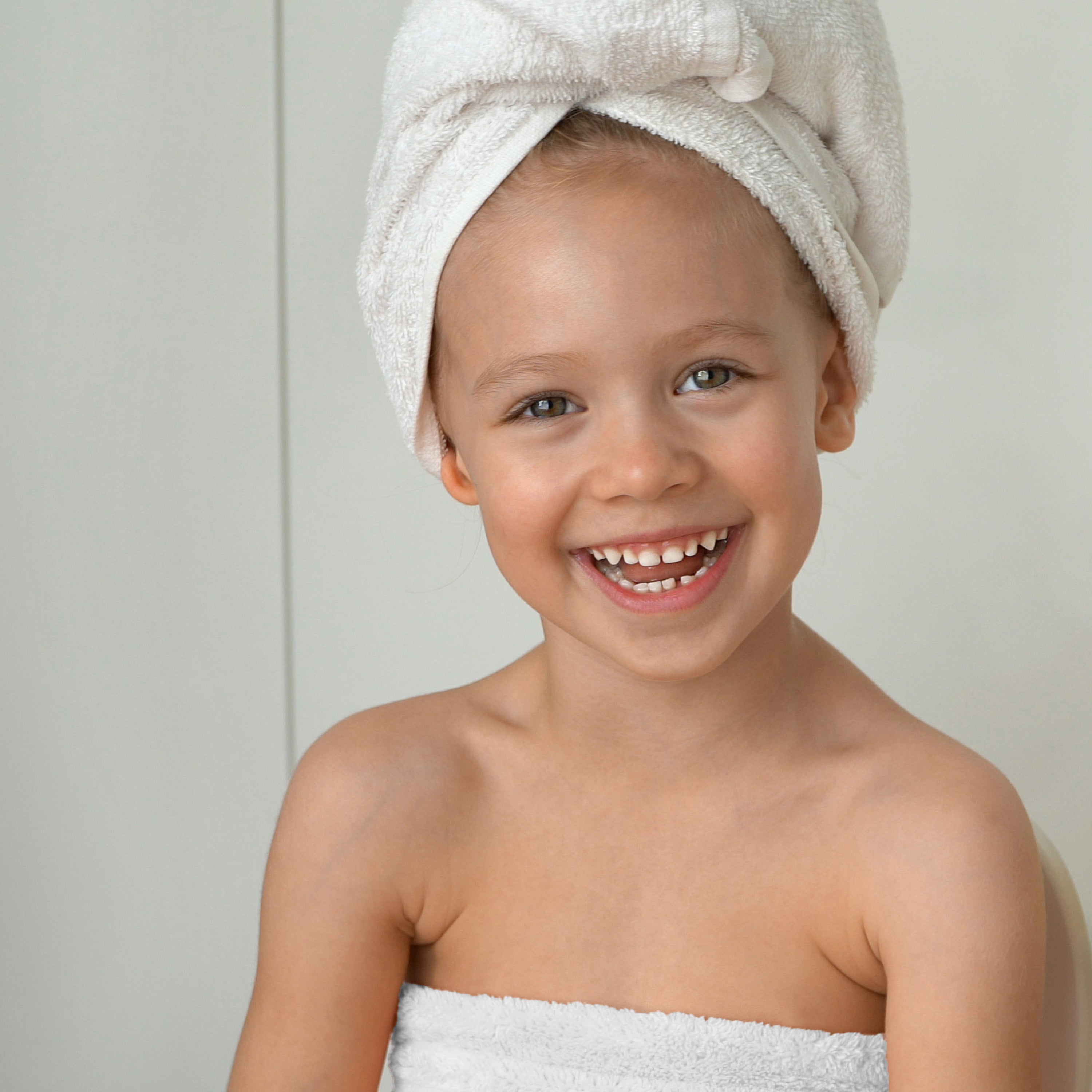 Child smiling while wearing a white towel wrap and a matching towel on their head, against a light background.