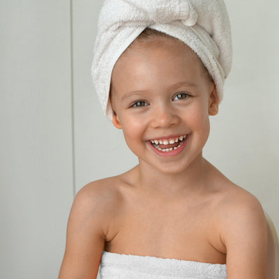Child smiling while wearing a white towel wrap and a matching towel on their head, against a light background.