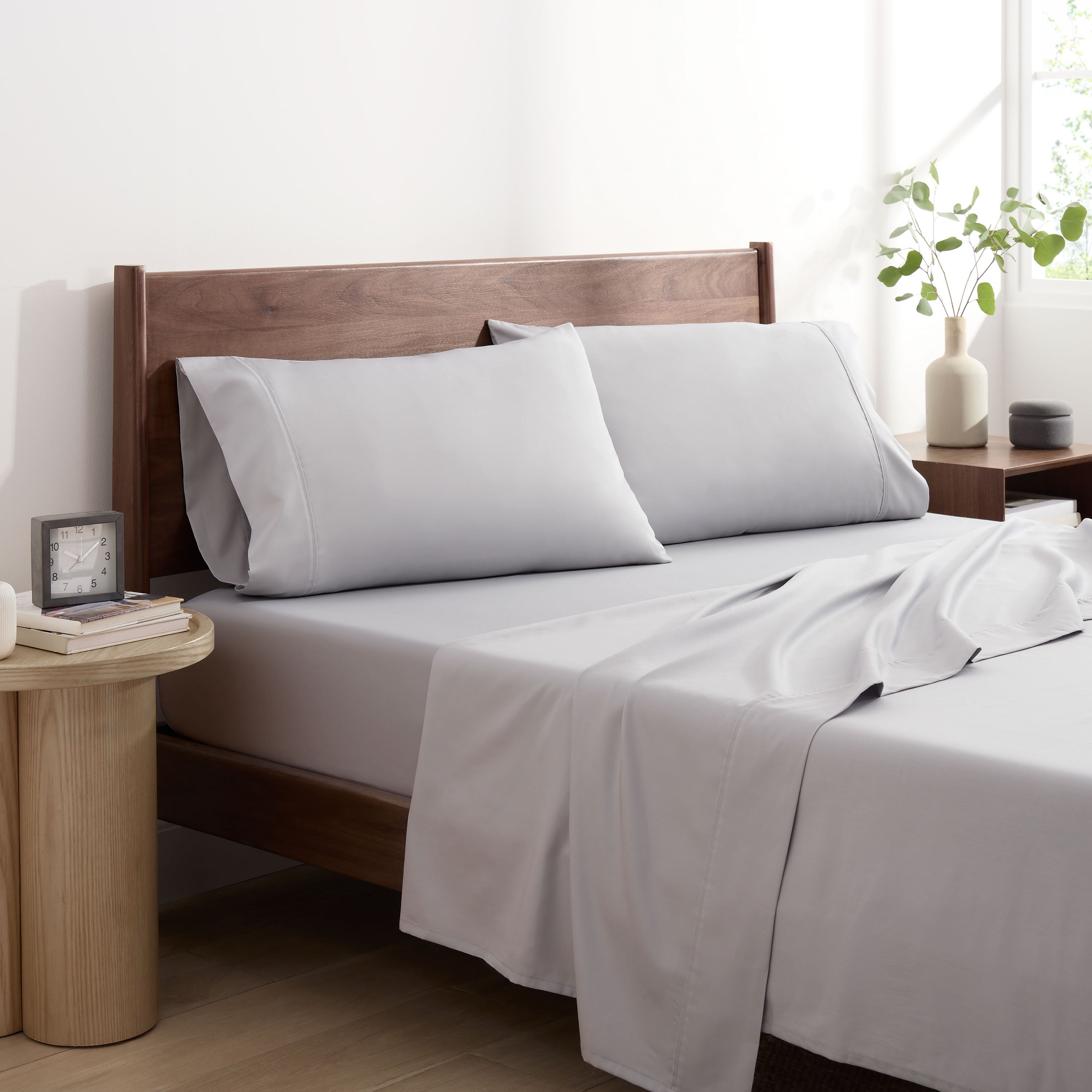 Light gray cotton and Tencel sheet set on a bed with two pillows, a nightstand, and a plant in the background.