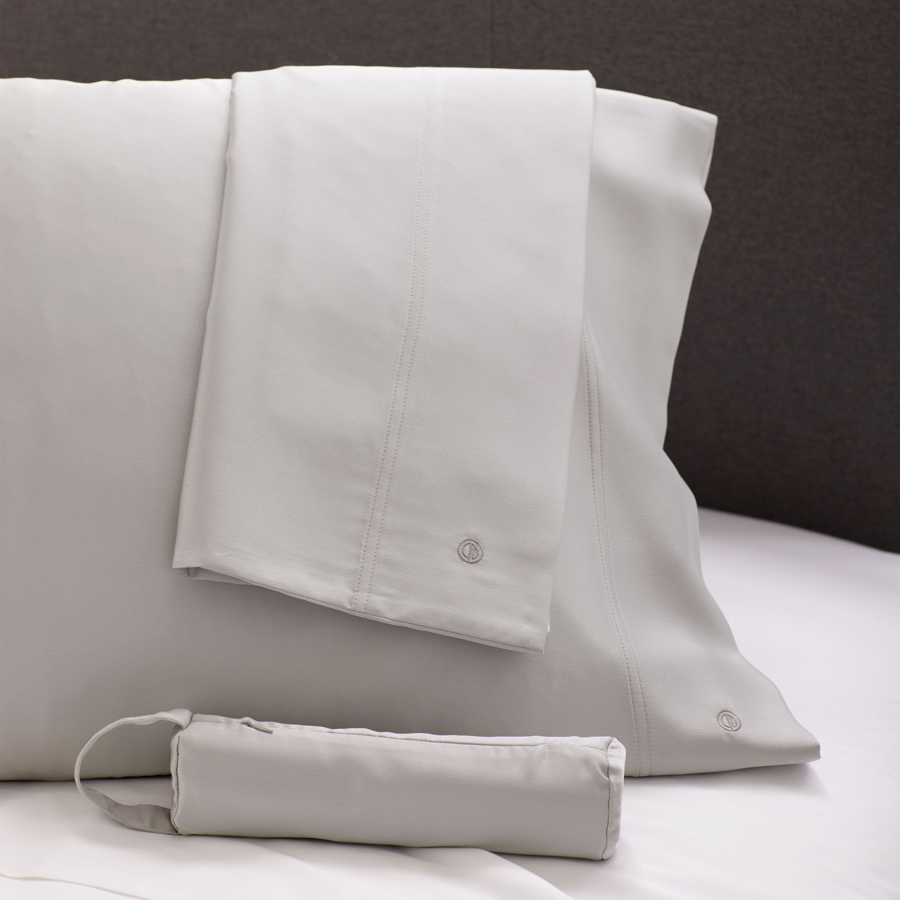 Satin standard pillowcase pair in light gray with a matching storage pouch, displayed on a bed.