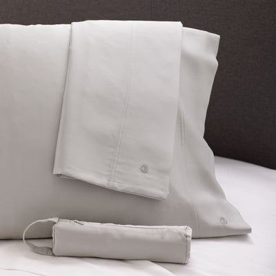 Satin standard pillowcase pair in light gray with a matching storage pouch, displayed on a bed.