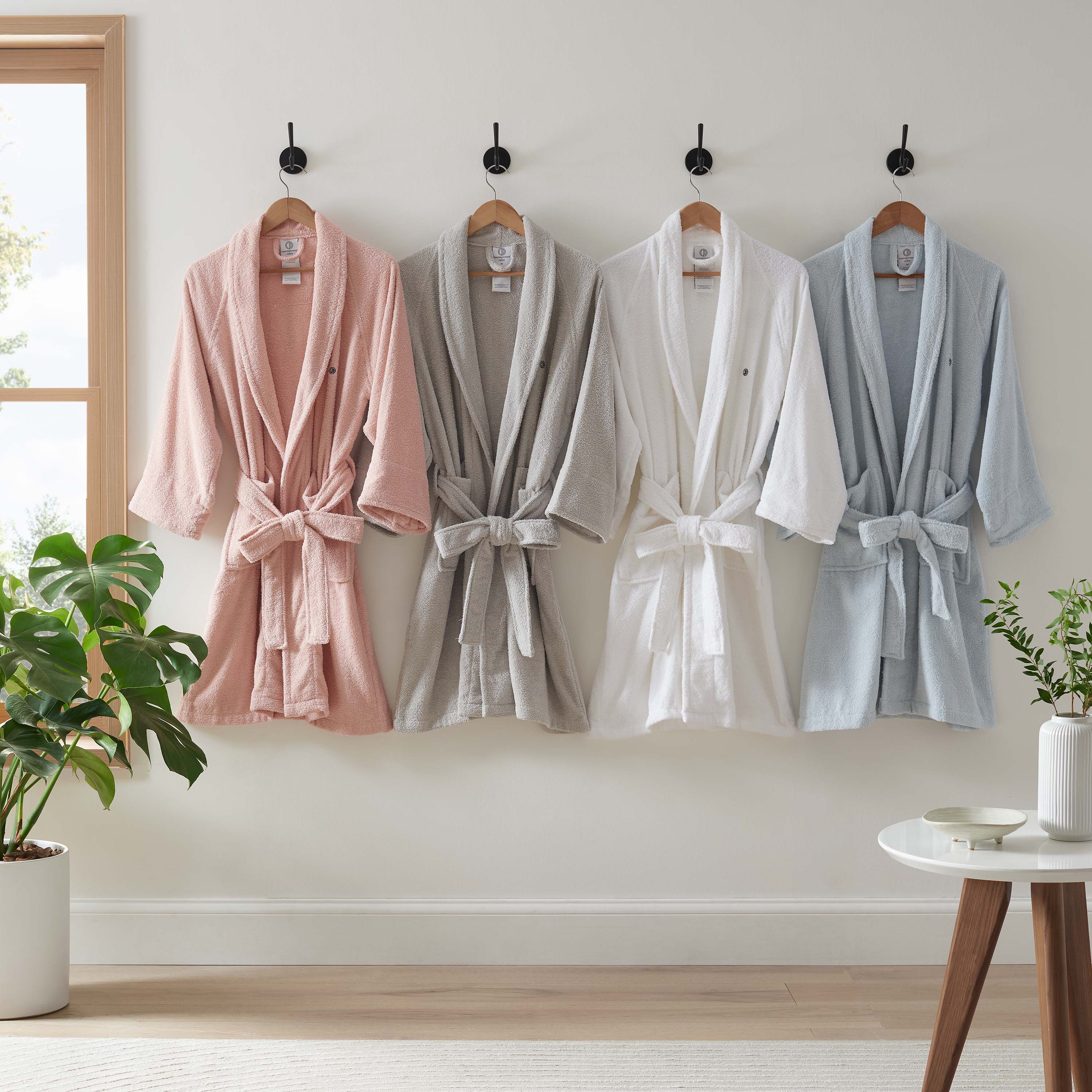 Four terry bath robes in pink, gray, white, and light blue hanging on hooks against a wall with a plant and table nearby.