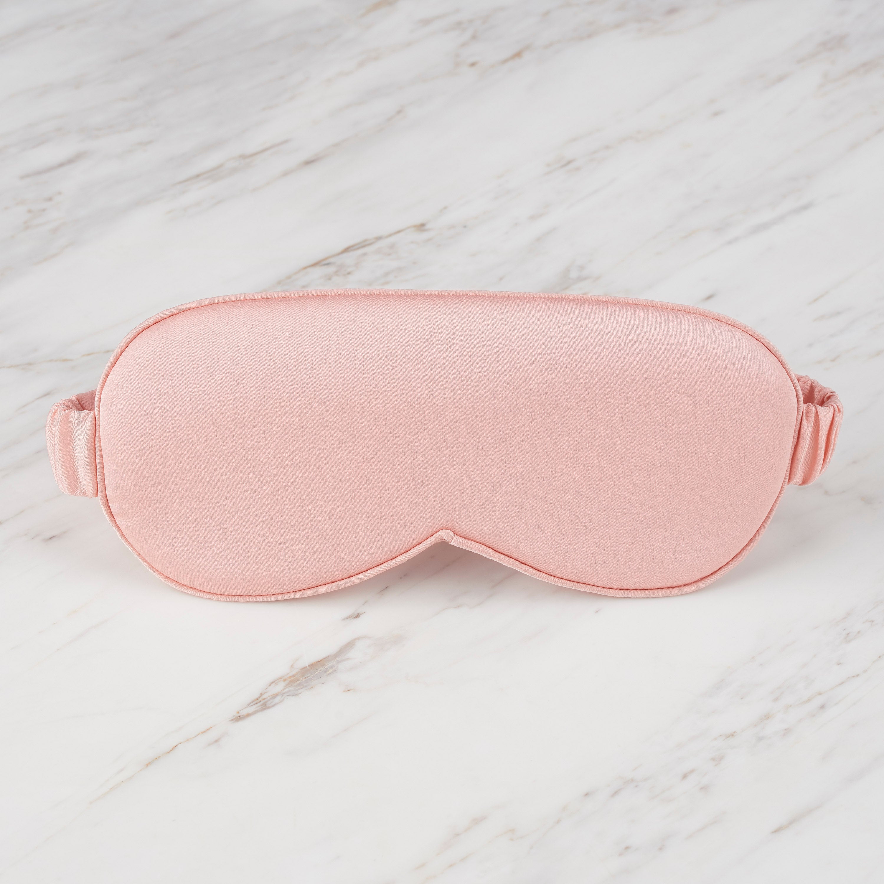 Pink satin eye mask with a soft texture and adjustable strap, placed on a marble surface.