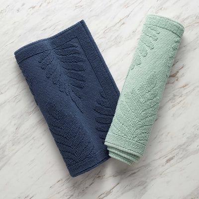 Two bath rugs in navy blue and light green with a fern pattern, displayed on a marble surface.