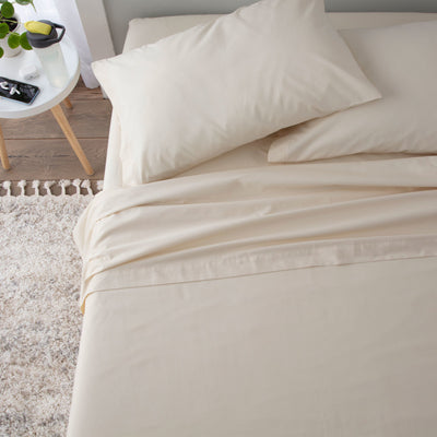 Beige sheet set with two pillows on a bed, alongside a small table with a plant and a water bottle.