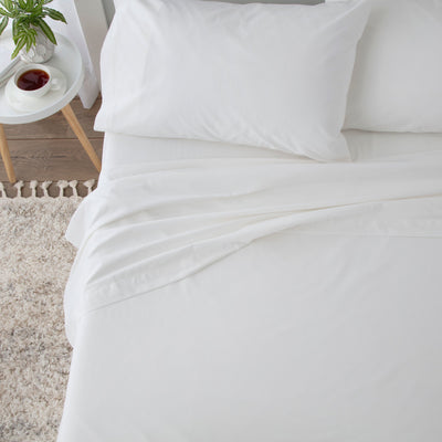 White sheet set with two pillows, draped over a bed, alongside a small table with a cup and plant.