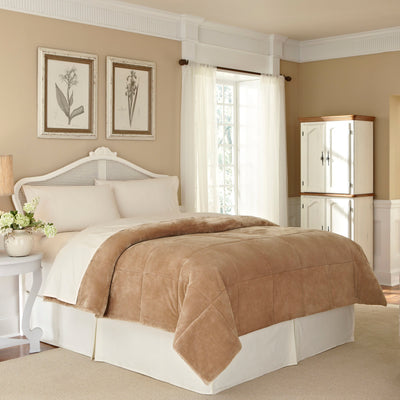 Tan plush blanket draped over a bed with white linens in a softly lit room featuring floral artwork and a cabinet.