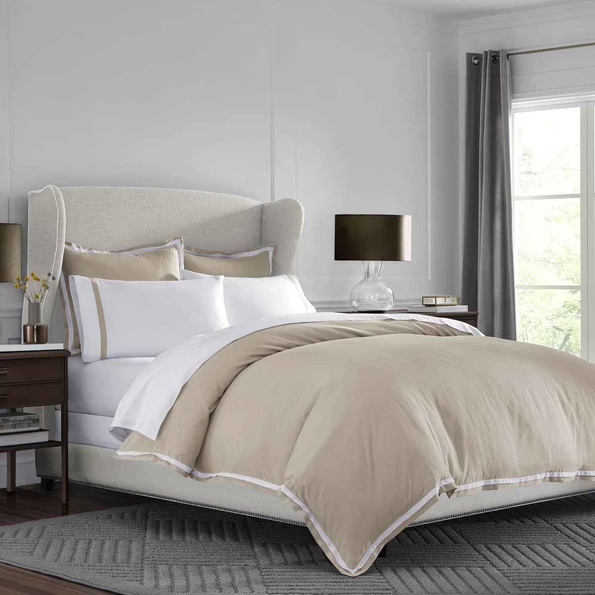 Duvet cover set in beige with white trim, paired with decorative pillows on a bed with a light gray rug.